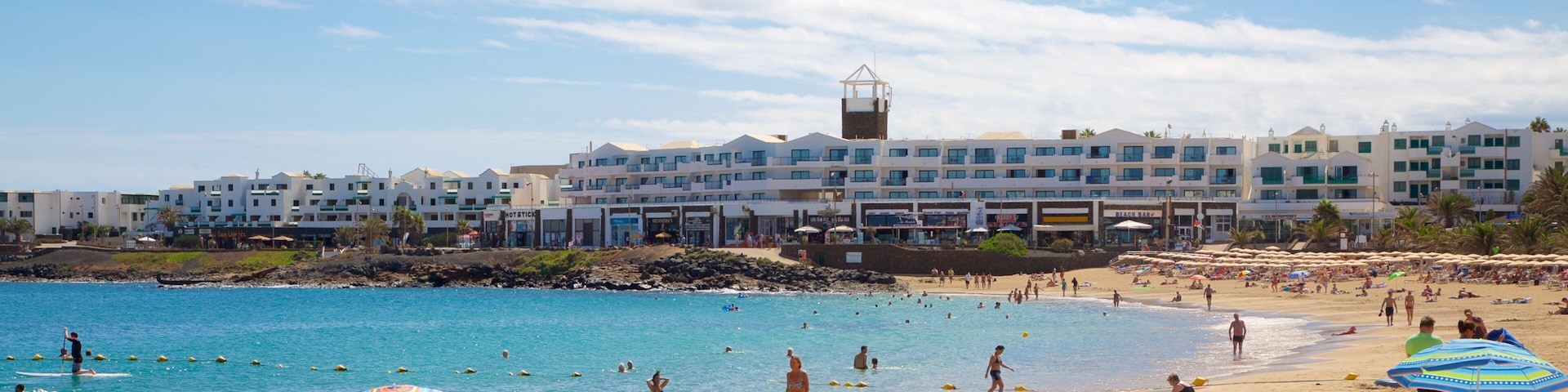A beach with people swimming and sunbathing, umbrellas, and buildings in the background.