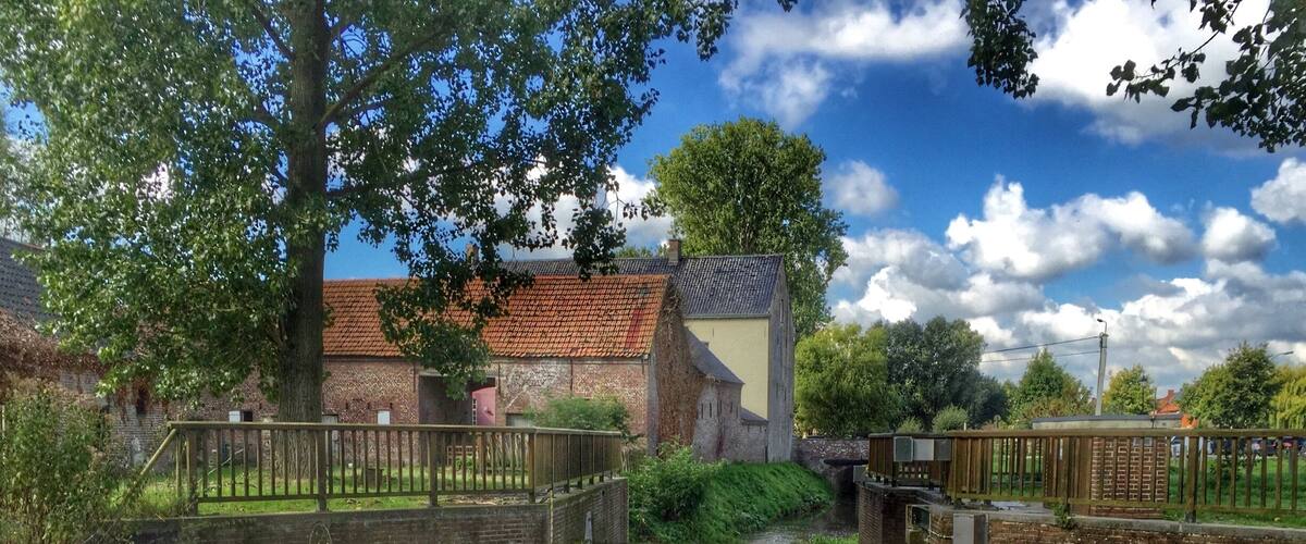 A calm canal with a wooden bridge, a stone building with a red roof, and lush greenery.