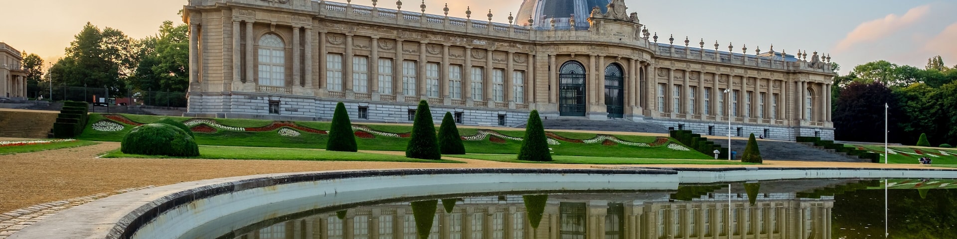 A grand building with a dome and columns, surrounded by a reflective pool and trimmed hedges.