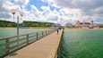 Een houten steiger die zich uitstrekt over een turquoise zee, met een heldere lucht en gebouwen op de achtergrond.