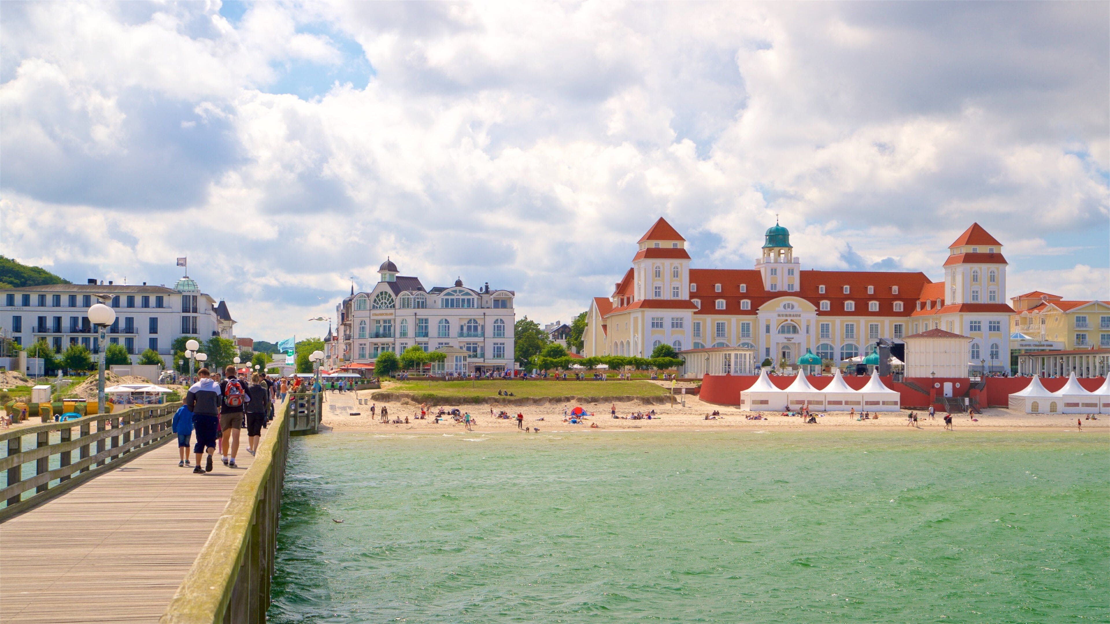 A pier with people walking, a beach with tents, and historic buildings with red roofs.