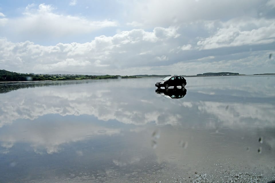 A car parked on a calm lake with a clear reflection of the sky and clouds.