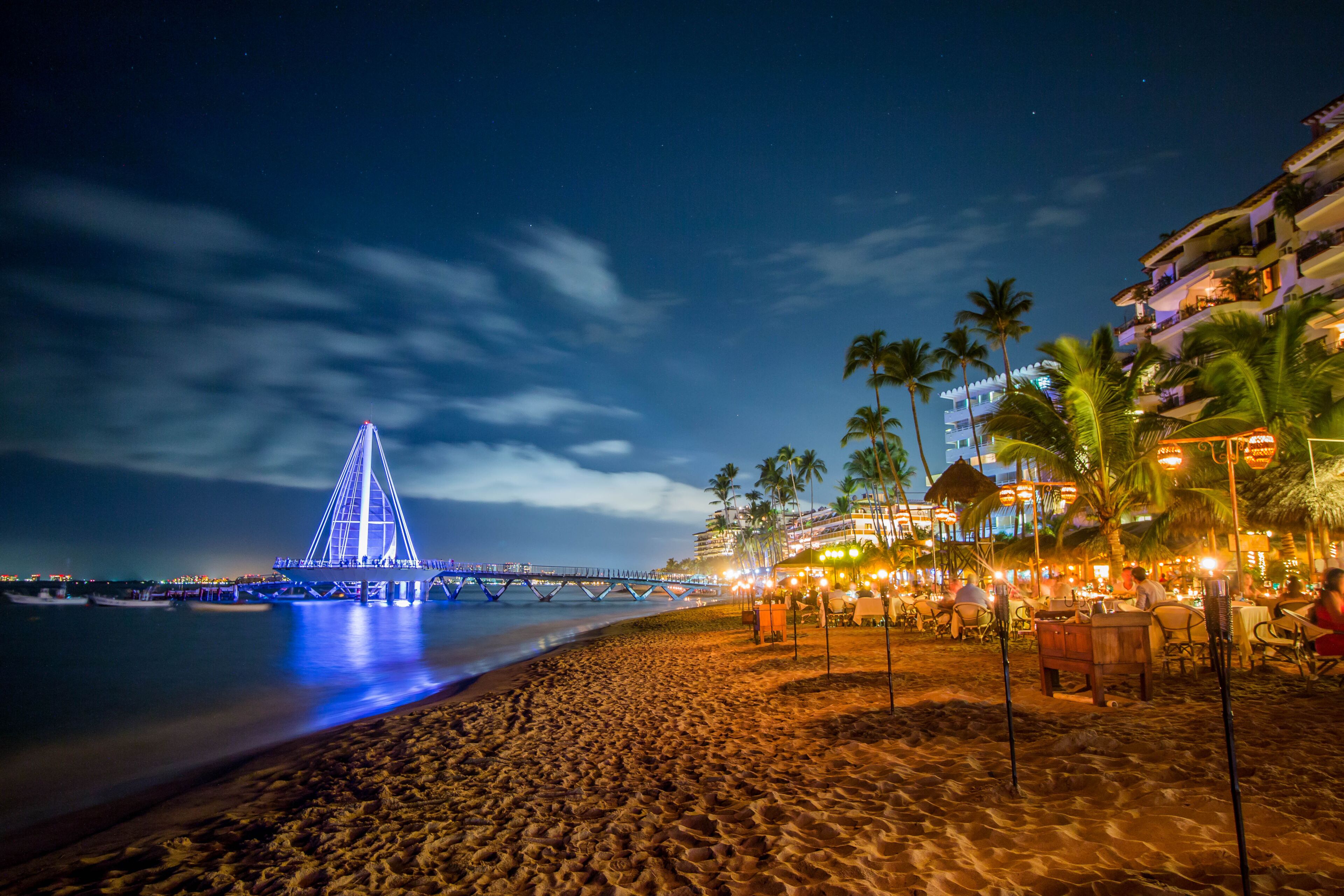 Une plage de nuit, avec un pont illuminé, des palmiers et un complexe hôtelier doté d’une terrasse.