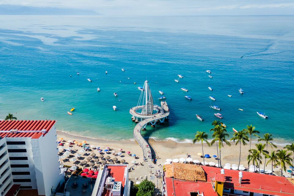 A coastal cityscape with a prominent pier, clear blue waters, and a beach area with umbrellas and palm trees.