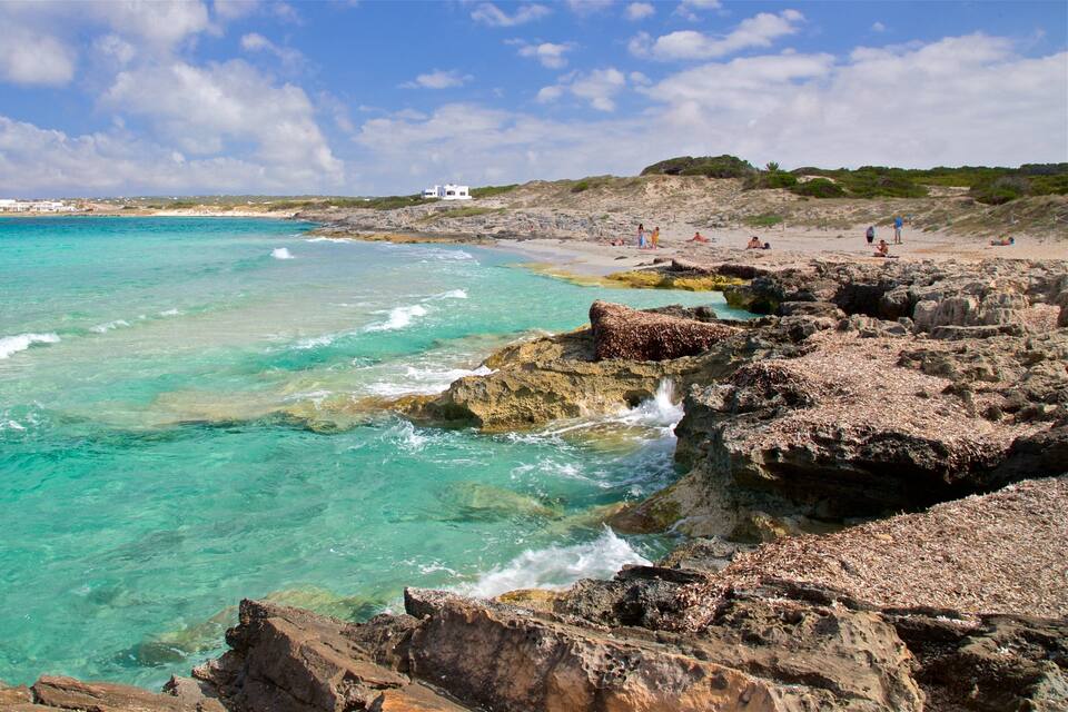 Eine felsige Küstenlinie mit klarem türkisblauem Wasser, einem Sandstrand und ein weißes Gebäude in der Ferne.