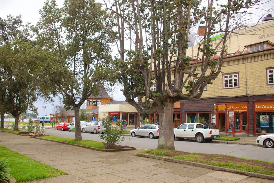 Una calle bordeada de árboles y coches aparcados, con edificios que incluyen un hotel y tiendas.
