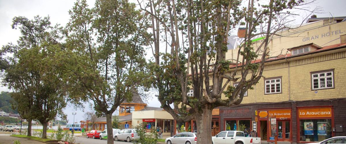 A street lined with trees and parked cars, with buildings including a hotel and shops.