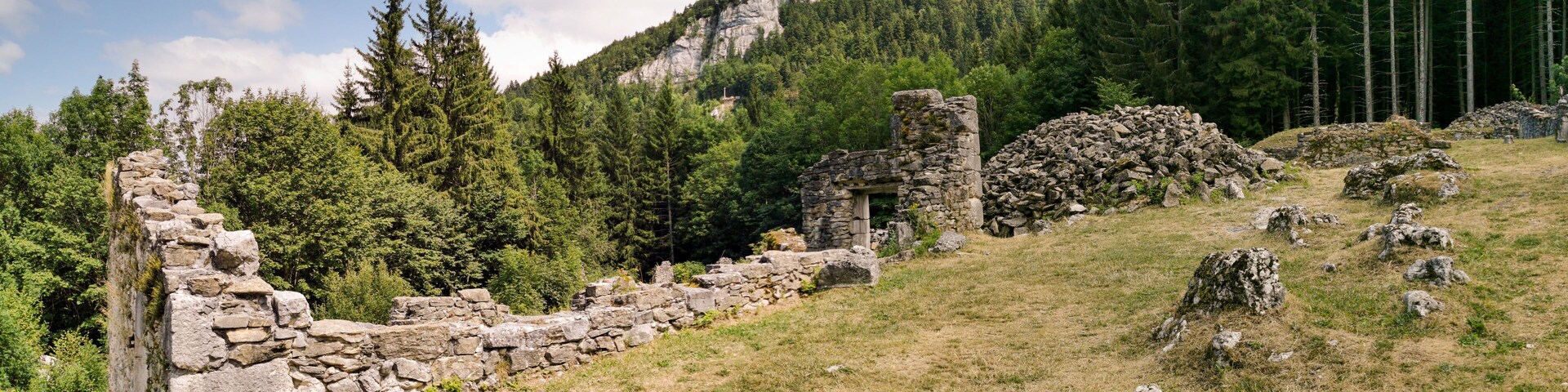 Antiguas ruinas de piedra en una ladera, con un bosque denso y una montaña al fondo.