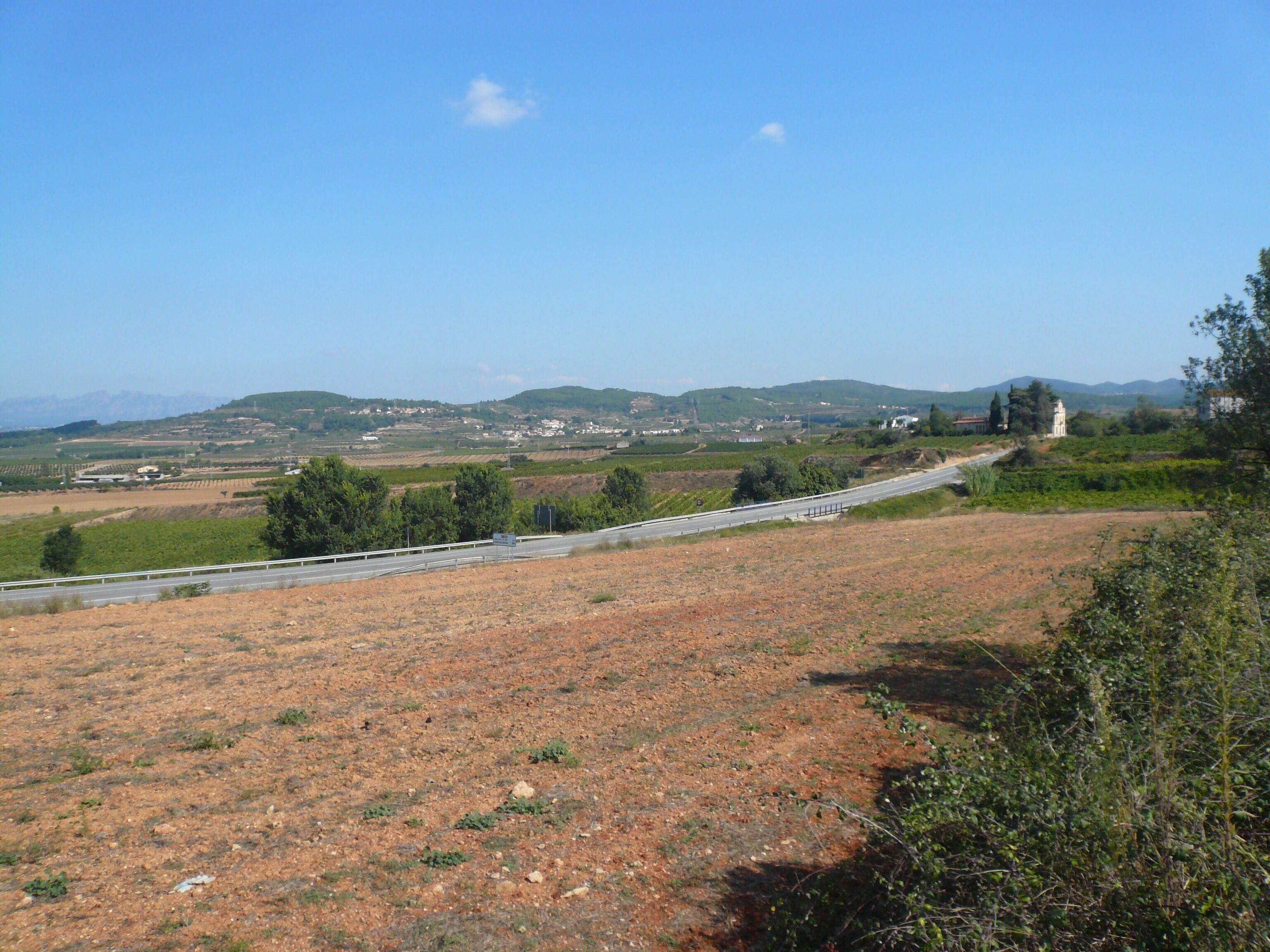 A rural landscape with a road curving through fields, distant hills, and scattered buildings.