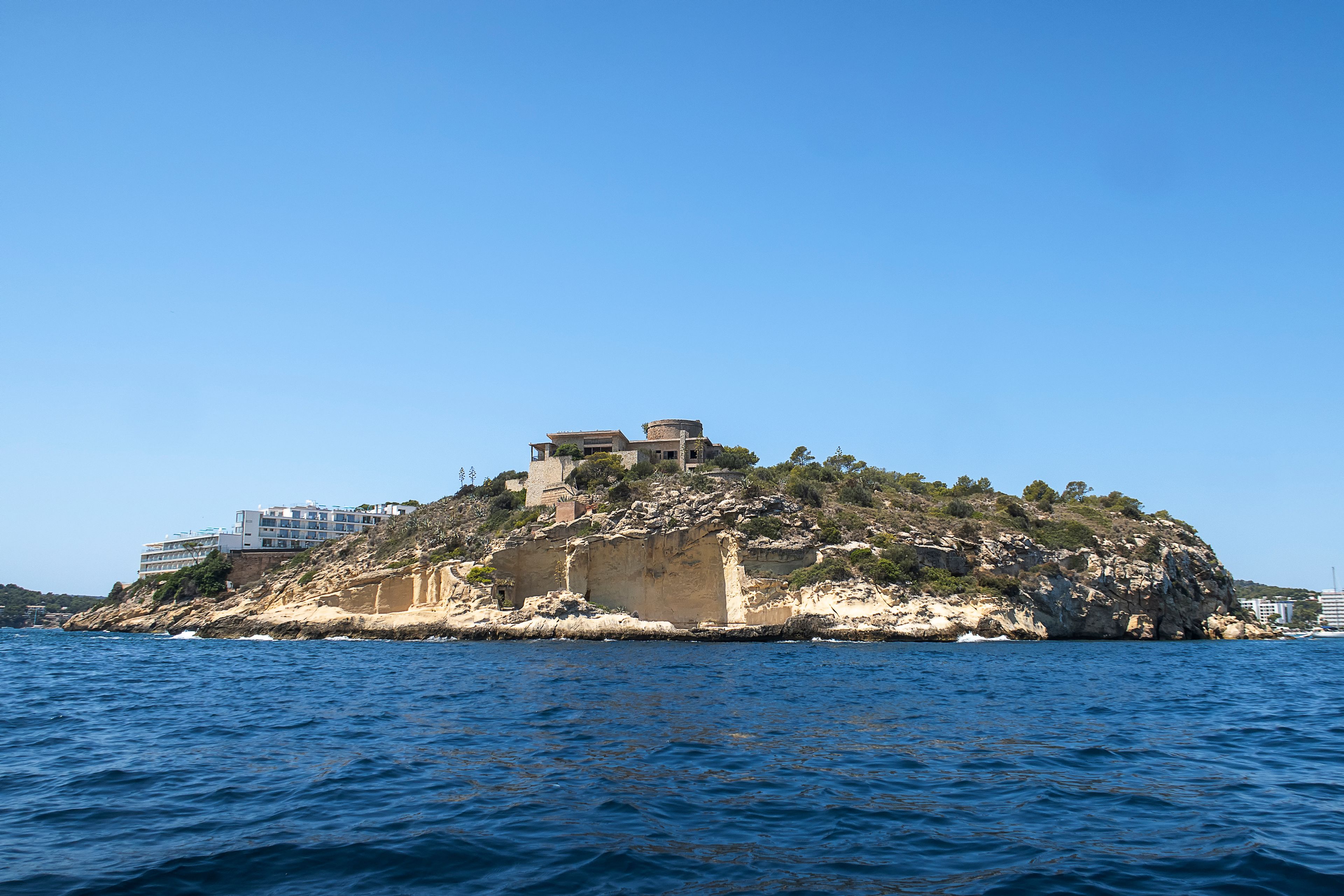 A rocky island with buildings on top, surrounded by clear blue water.