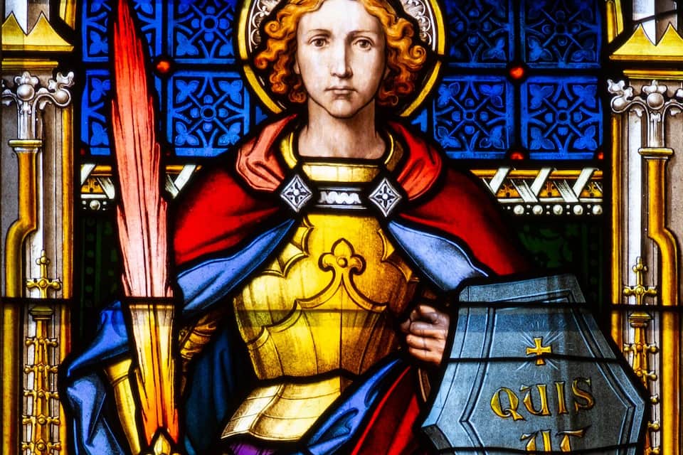 A stained glass window with a figure holding a sword and shield, surrounded by ornate architectural details.