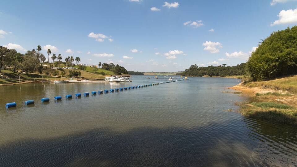 A river with blue floating barriers, boats, and a grassy bank with trees.