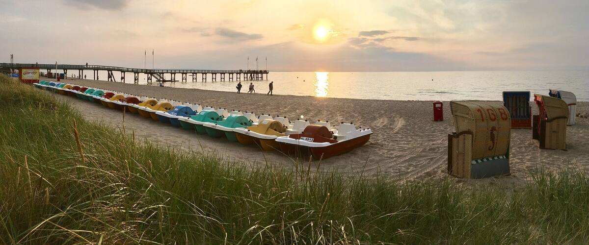 A beach with a row of colorful boats lined up on the sand, a pier extending into the water, and beach huts in the foreground.