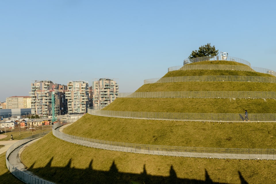 Uma colina gramada em terraços com uma cerca, uma árvore e edifícios modernos ao fundo.