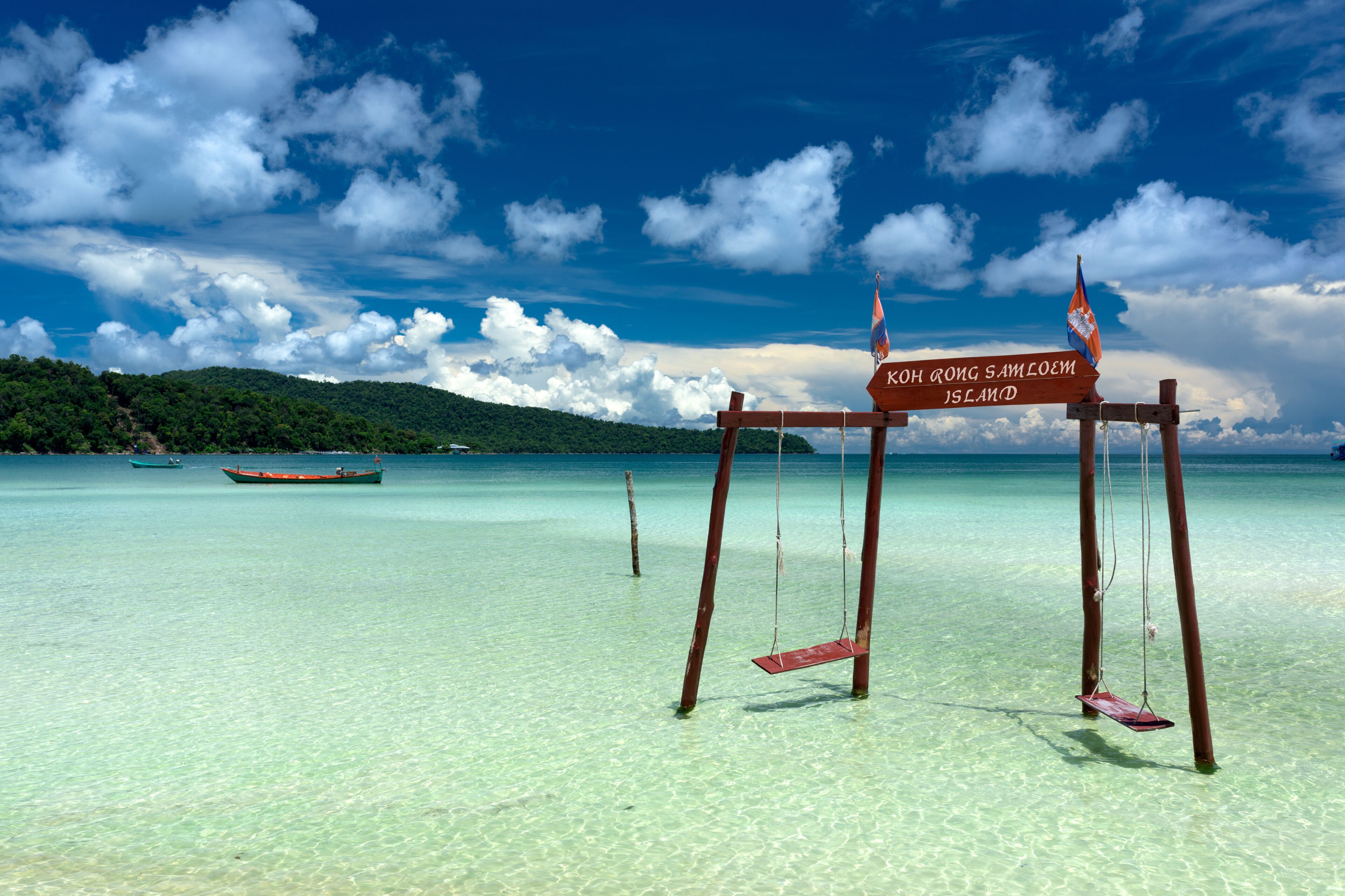 A clear turquoise sea, a wooden swing set, a boat, and a sign with 'Koh Rong Samloem Island' written on it.