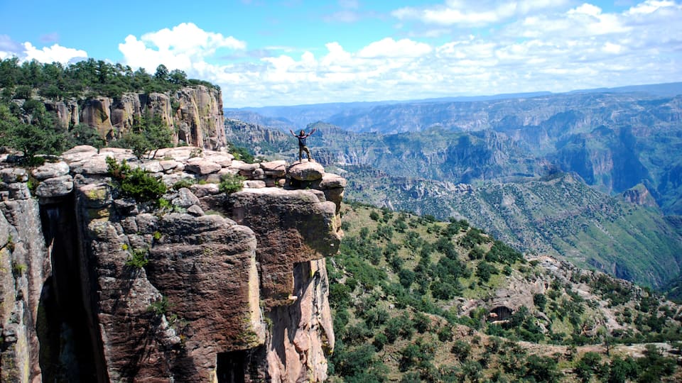 A person standing on a rocky cliff with a vast canyon landscape in the background.