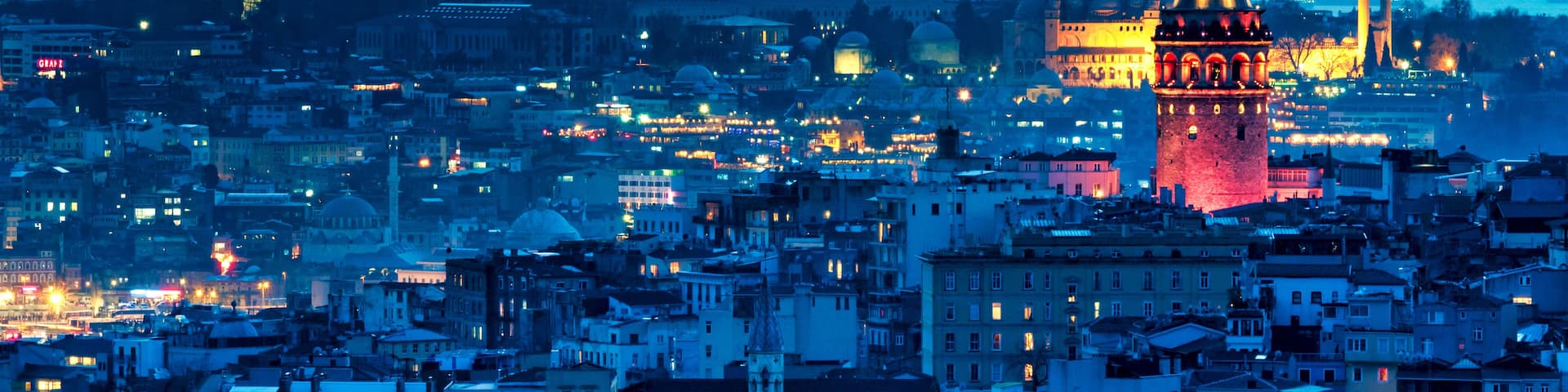 A cityscape at dusk with prominent towers and domes, illuminated buildings, and a dense urban layout.