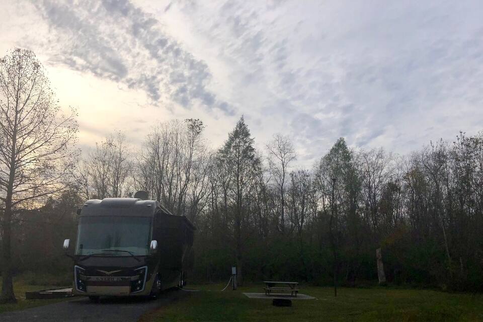 Una autocaravana gran aparcada en una zona boscosa, amb una taula de picnic i una bomba d'aigua al fons.
