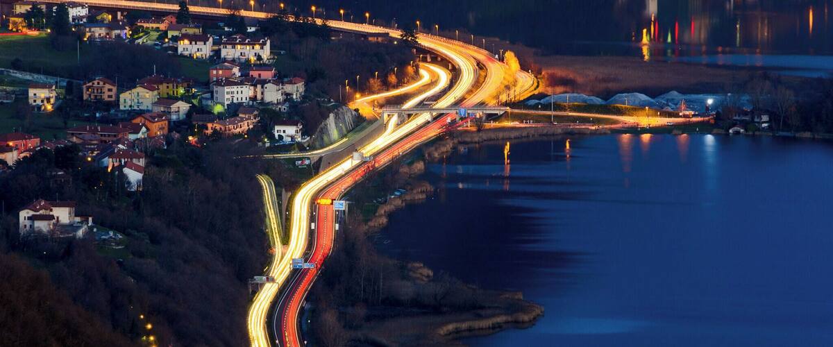 A nighttime aerial view of a winding road along a lakeside with buildings and a bridge in the background.