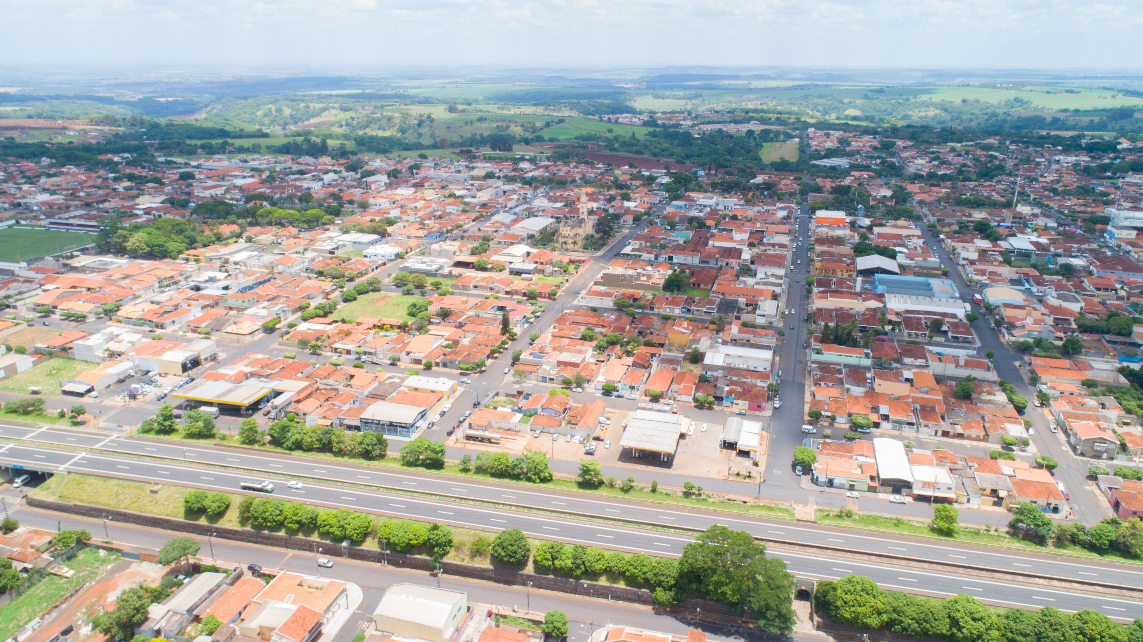 An aerial view of a town with red-roofed buildings, a main road, and surrounding green fields.
