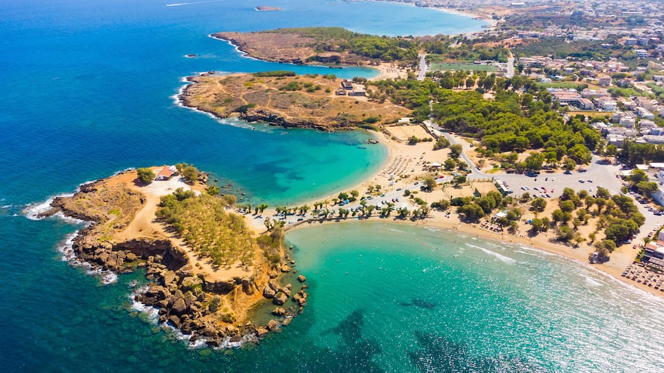 An aerial view of a coastal town with clear blue waters, a sandy beach, and a rocky peninsula.