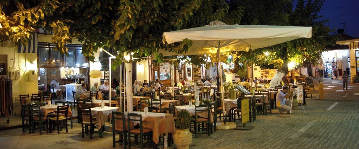 An outdoor café with tables and chairs under umbrellas, surrounded by trees and buildings.