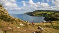 A hiker on a grassy hillside overlooking a coastal landscape with rolling hills and a body of water.