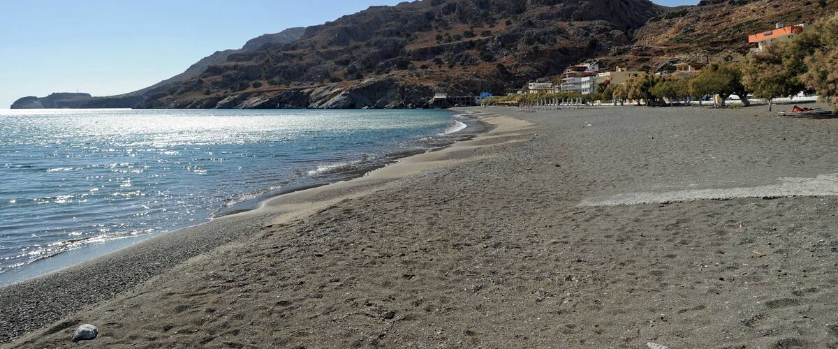 A pebbly beach with a mountain in the background and buildings along the shore.