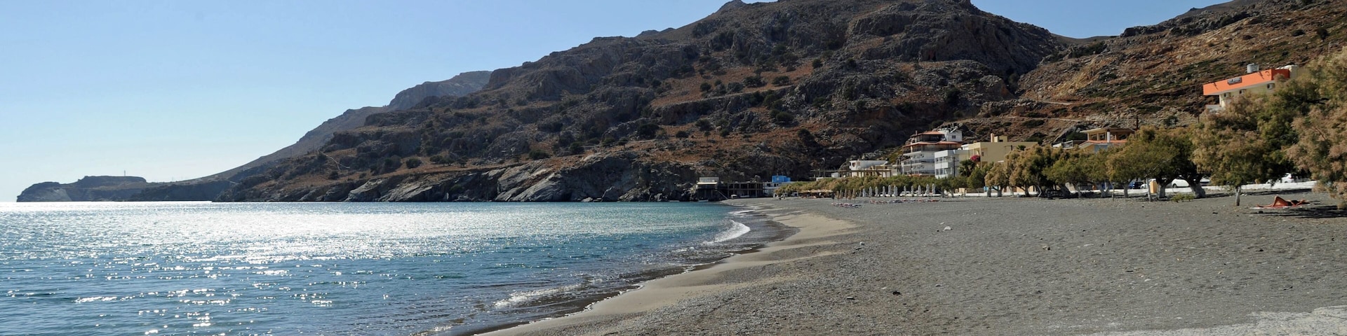 A pebbly beach with a mountain in the background and buildings along the shore.