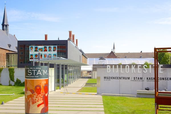 A museum entrance with a modern glass canopy, a historic building with a spire, and a large sign for the Stadsmuseum Gent.