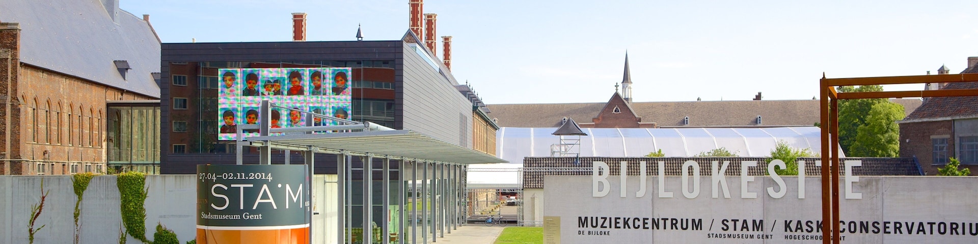 Ein Museumseingang mit moderner Glasüberdachung, ein historisches Gebäude mit Kirchturm und ein großes Schild für das Stadsmuseum Gent.