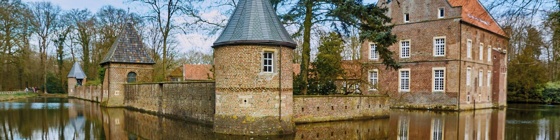 A historic building with a tower and a large brick house by a calm lake.