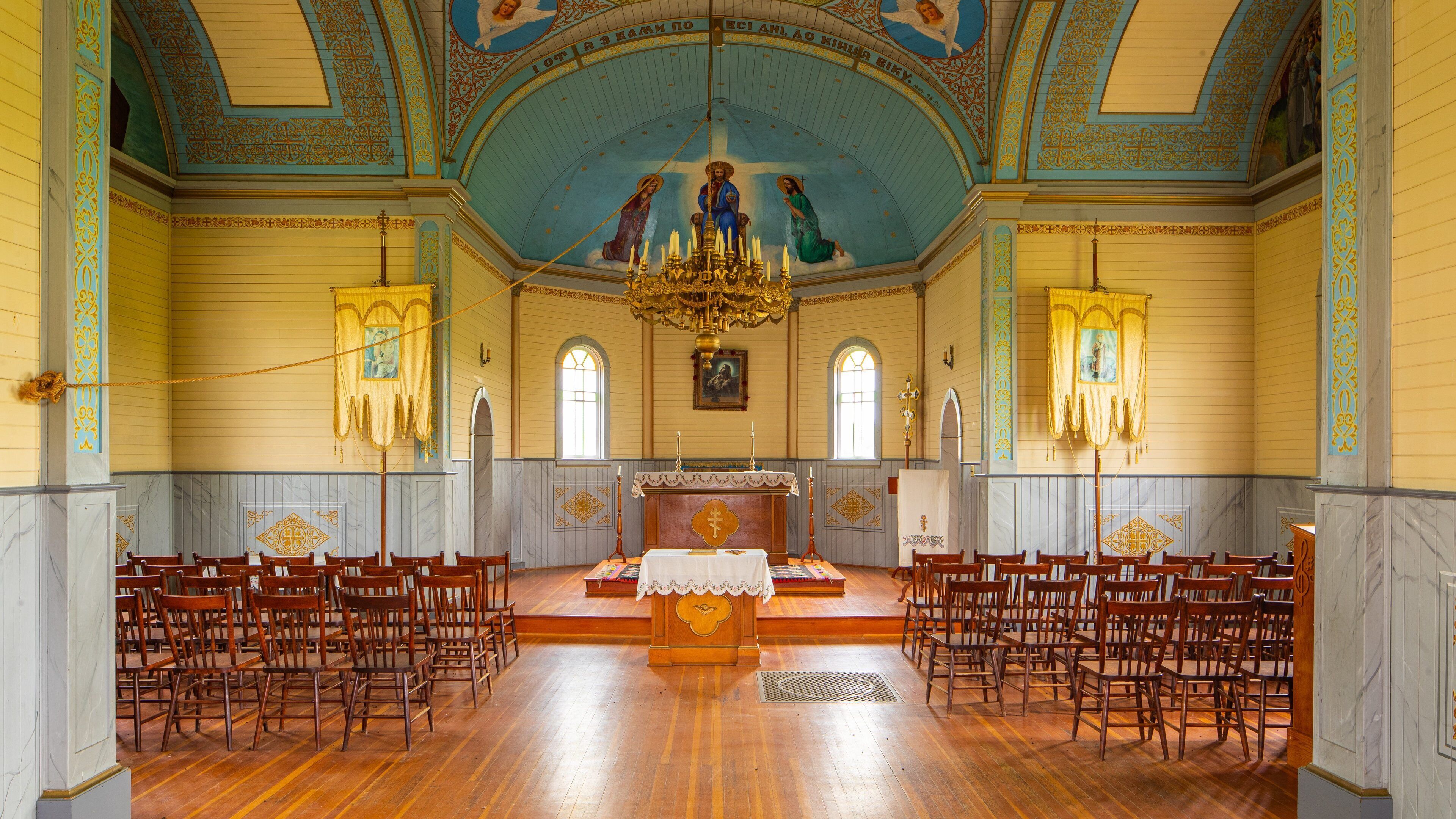 A church interior with wooden pews, a central altar, and a blue ceiling with religious artwork.