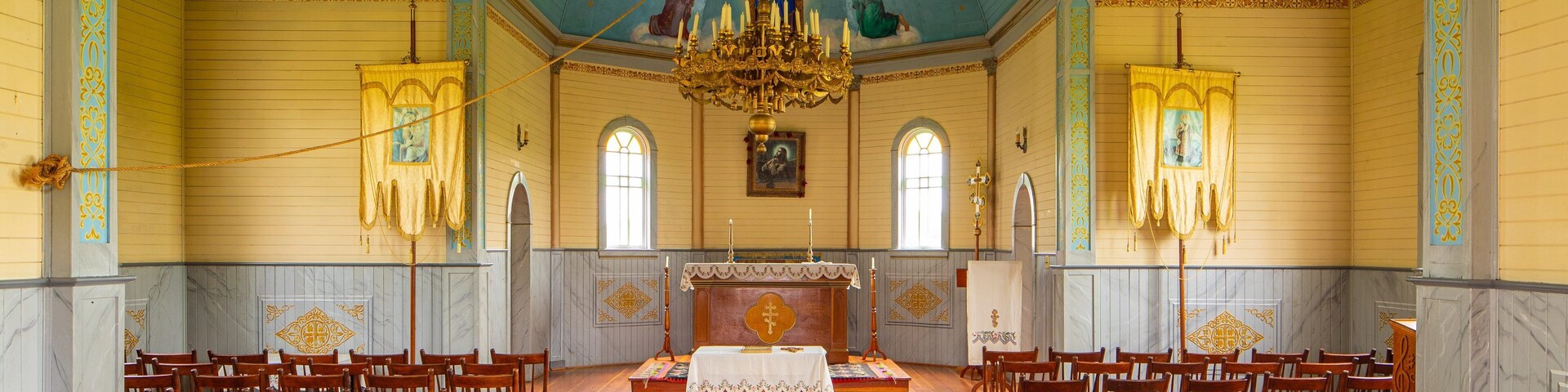 A church interior with wooden pews, a central altar, and a blue ceiling with religious artwork.