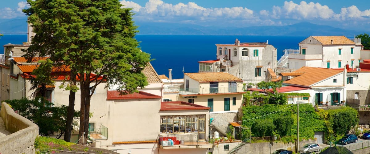 A coastal town with Mediterranean architecture, a prominent tree, and the sea in the background.
