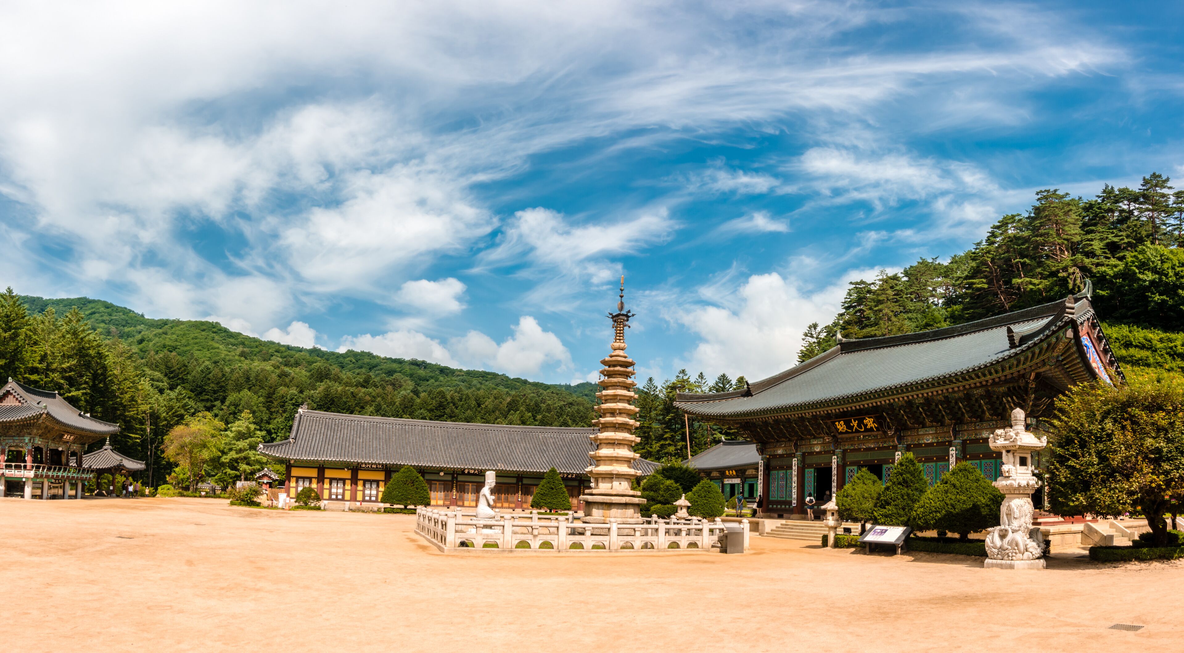 A traditional Korean temple complex with pagodas, a central stone lantern, and lush green mountains in the background.