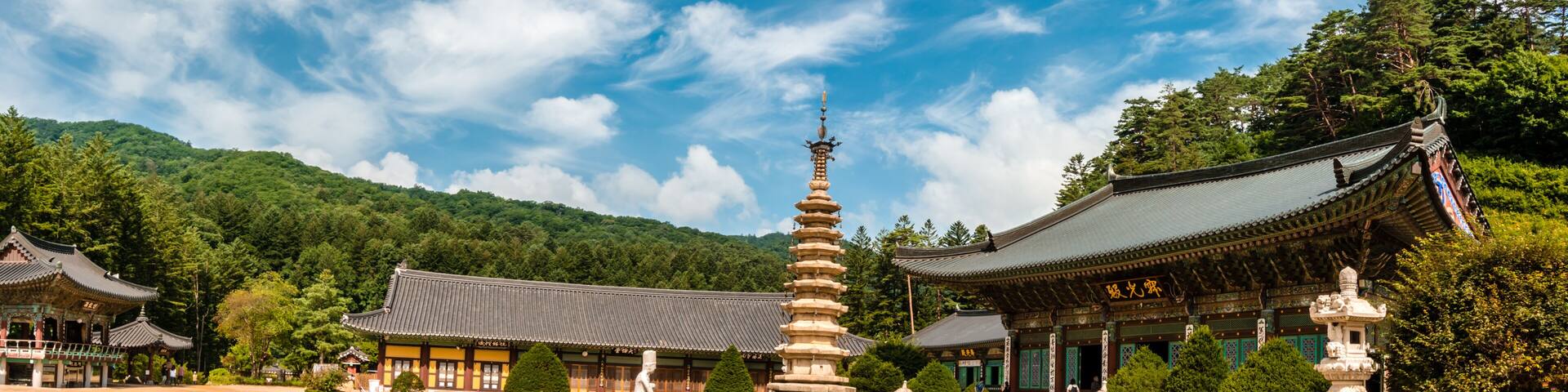 A traditional Korean temple complex with pagodas, a central stone lantern, and lush green mountains in the background.