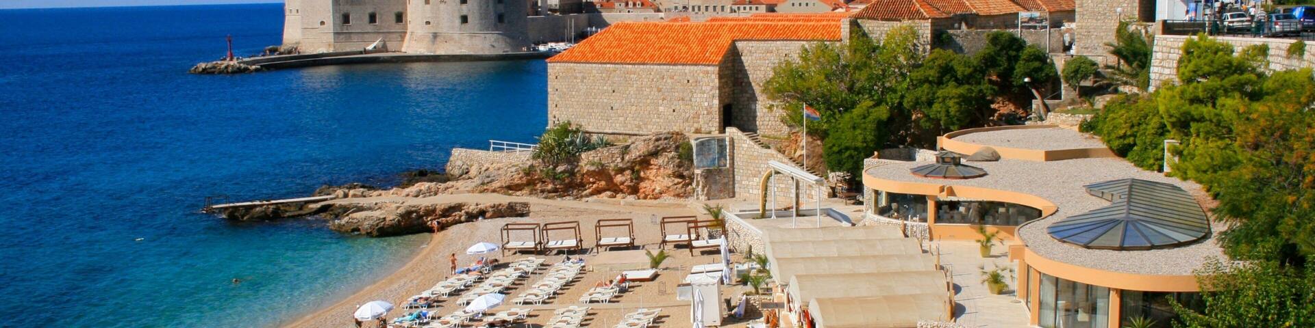A coastal city with a historic fort, a sandy beach with sun loungers, and a clear blue sea.