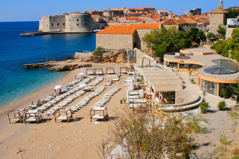 A coastal city with a historic fort, a sandy beach with sun loungers, and a clear blue sea.
