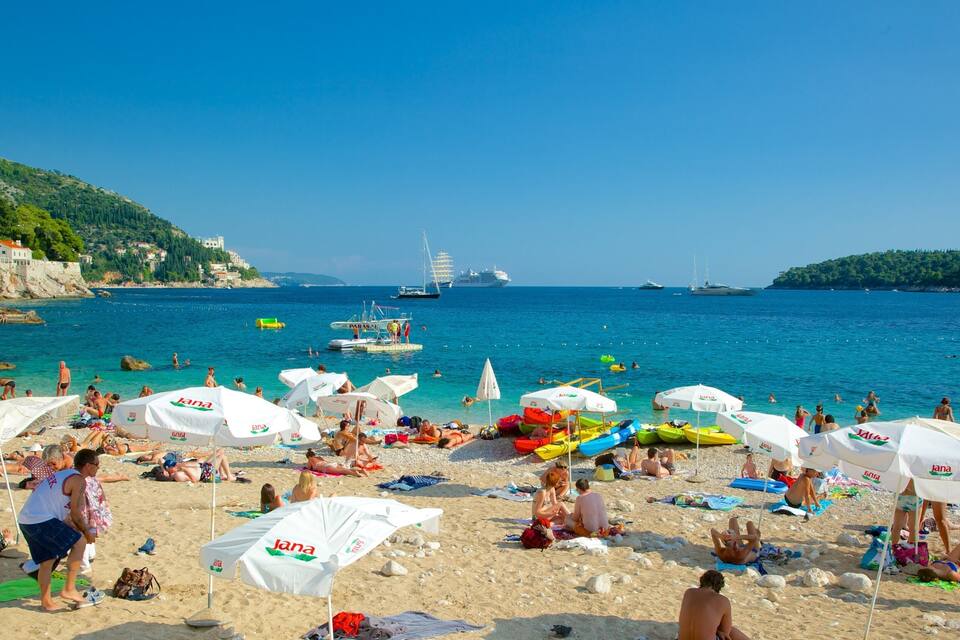 A crowded beach with people sunbathing and swimming, surrounded by clear blue waters and boats.