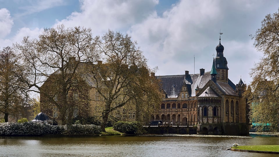A historic building with a tower, surrounded by trees and a body of water.
