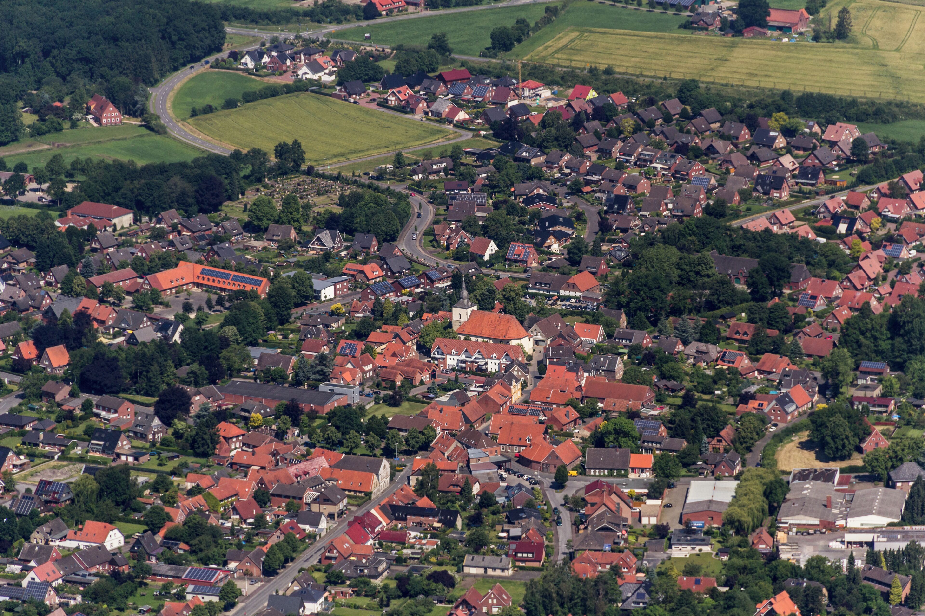 An aerial view of a densely populated village with red-roofed houses, green fields, and a winding road.