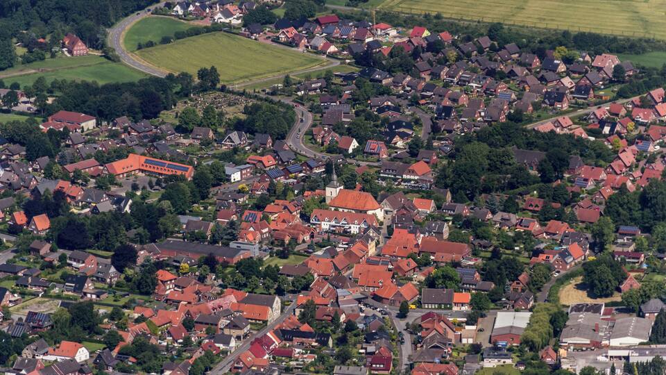 An aerial view of a densely populated village with red-roofed houses, green fields, and a winding road.