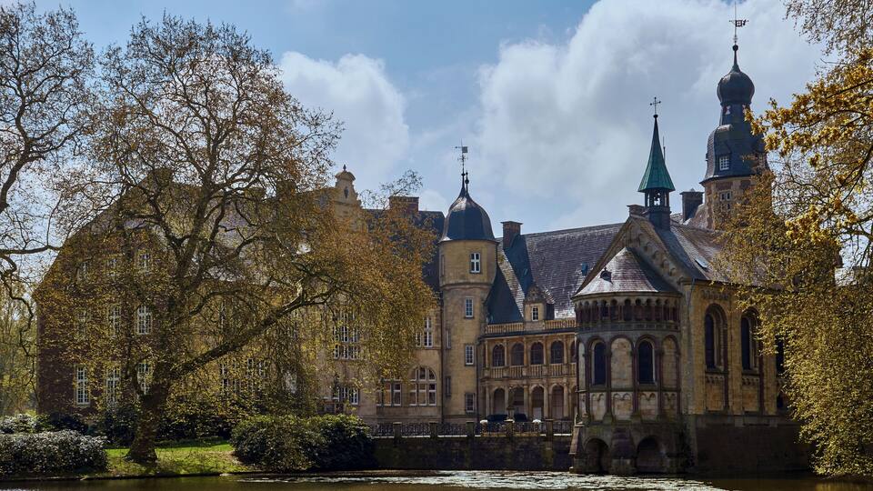 A historic building with multiple towers and domes, surrounded by a body of water and autumn foliage.