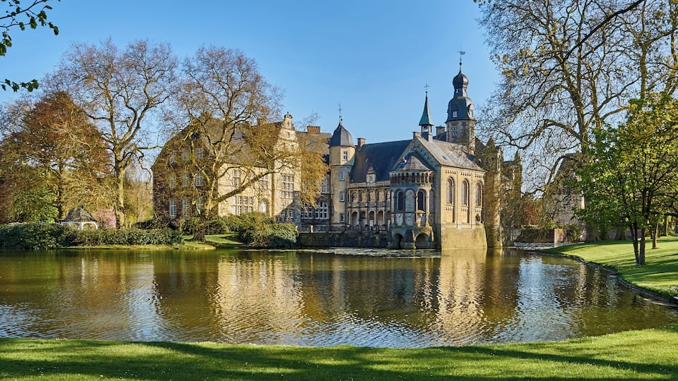 A historic building with a large pond in front, surrounded by trees and a well-maintained lawn.