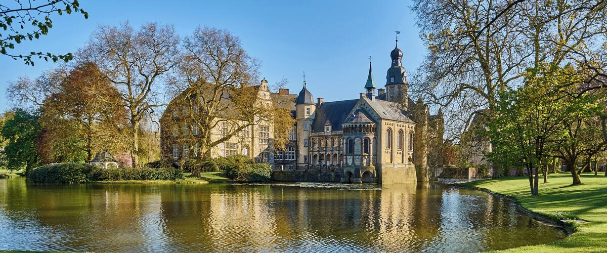 A historic building with a large pond in front, surrounded by trees and a well-maintained lawn.