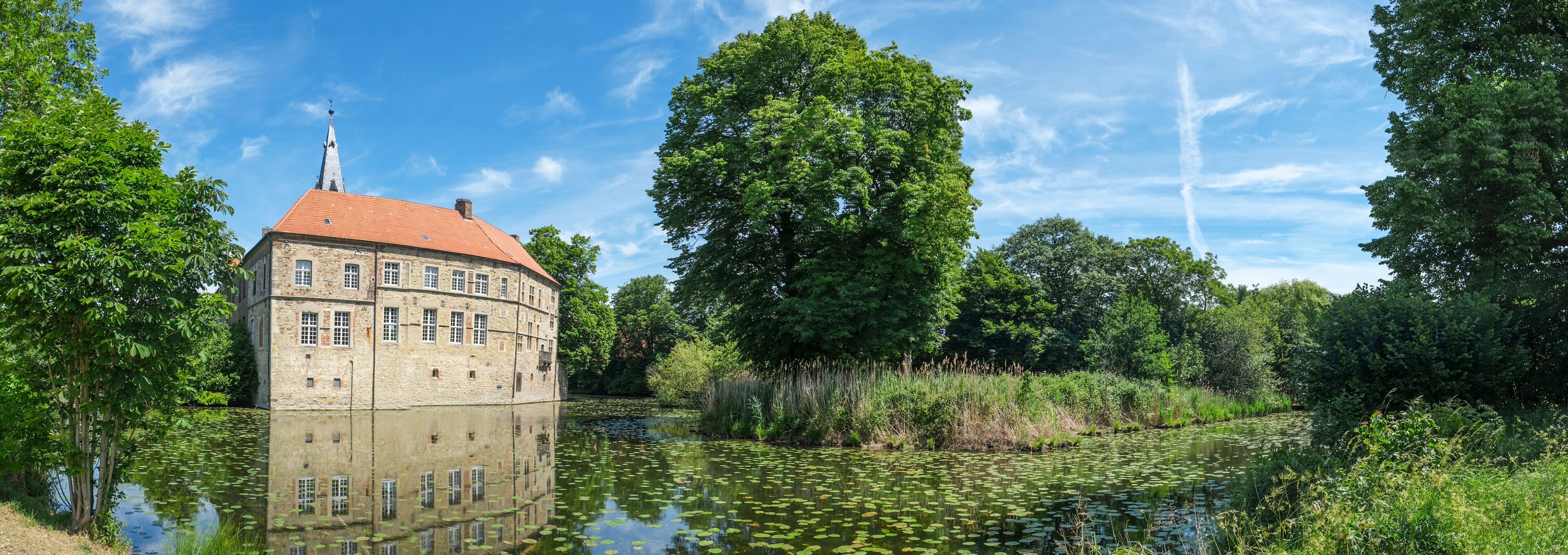 A historic building with a red roof and stone walls, surrounded by lush greenery and a calm pond with lily pads.