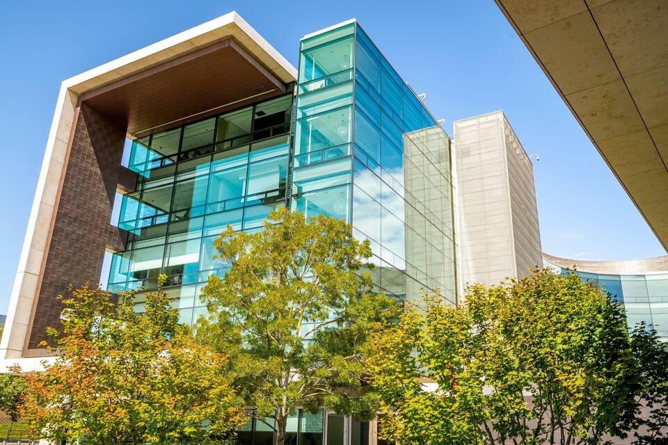 A modern glass building with a unique architectural design, surrounded by trees and a clear blue sky.