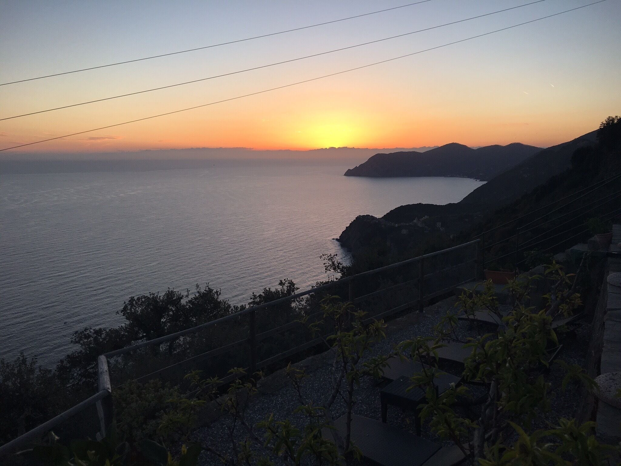 A sunset over a calm sea with rocky coastline and a wooden fence in the foreground.