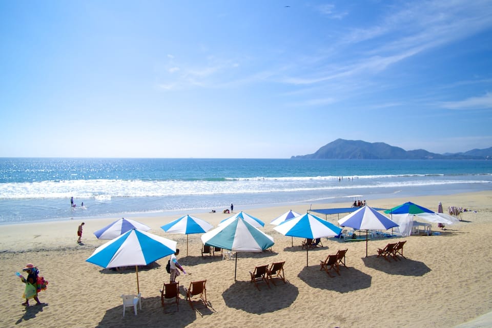 A beach with blue and white umbrellas, people, and a mountain in the background.